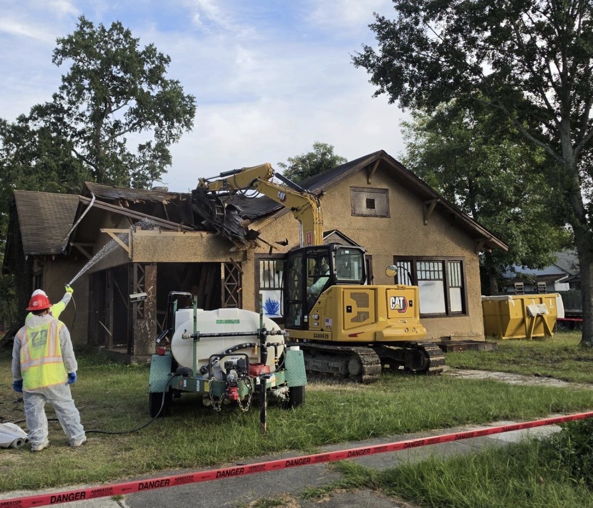 A-1 crew demolishing a house with an excavator while a worker in a protective suit wets down the debris behind asbestos warning tape