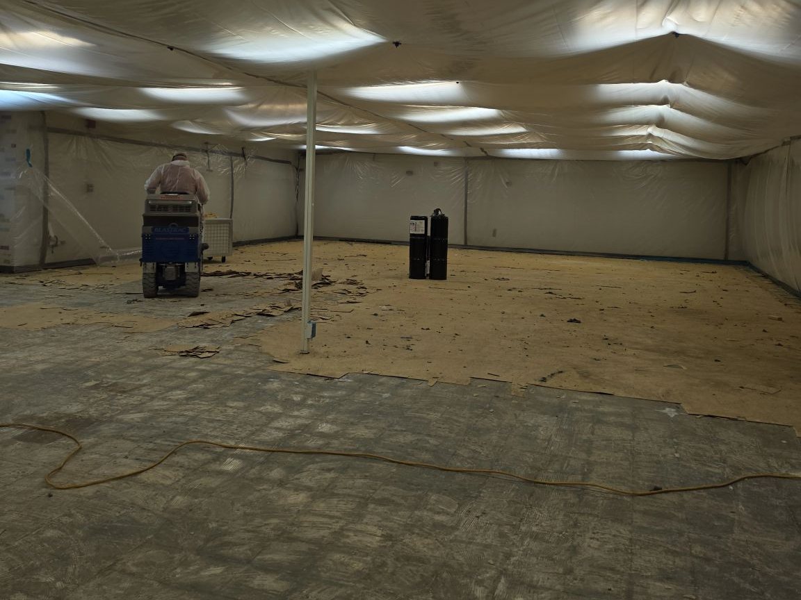 Crew member in a protective suit running a ride-on floor scraper inside an asbestos containment