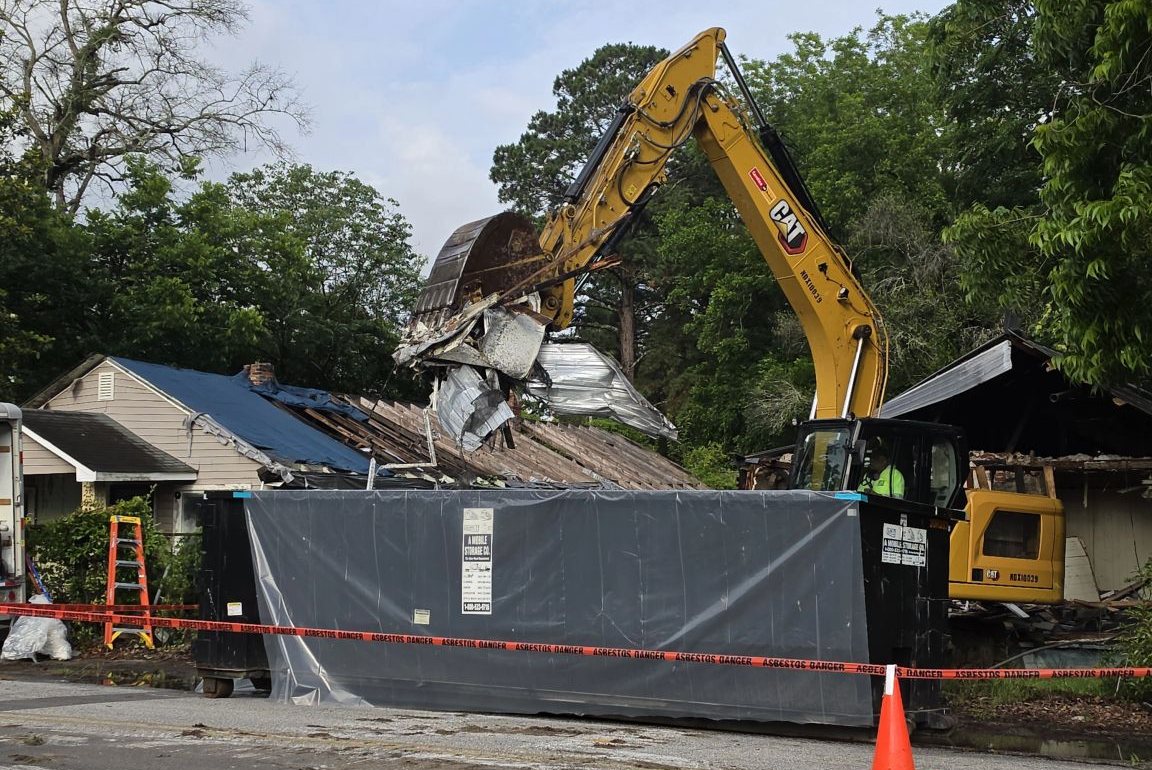 Excavator loading demolition debris into a lined, covered container behind asbestos warning tape