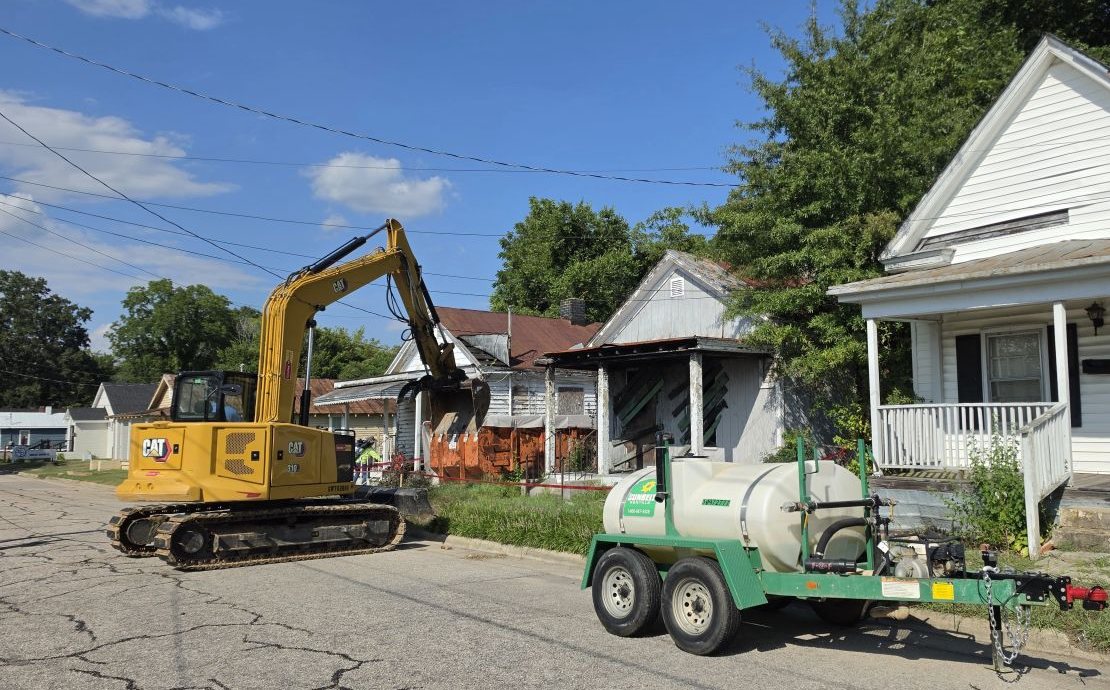 Excavator taking down a house on a neighborhood street with a water trailer standing by